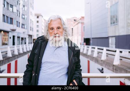 Serious bearded man with gray hairs wearing casual outfit and looking into camera while leaning on metal railing of modern bridge against downtown Stock Photo
