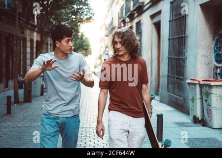 Carefree interested multiethnic men in casual clothes with longboard gesturing and talking while strolling along city street Stock Photo