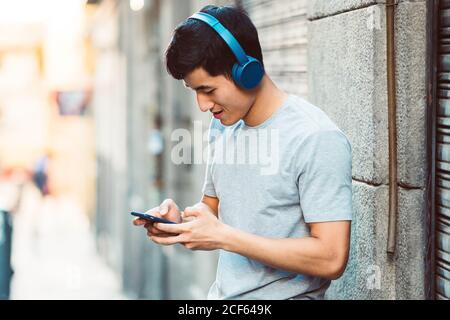 Joyful casual ethnic man in headphones using smartphone while standing on sunny city street Stock Photo