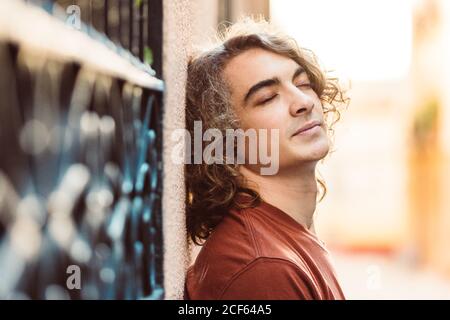 Side view of content relaxed man leaning on wall with closed eyes while standing on sunny street Stock Photo