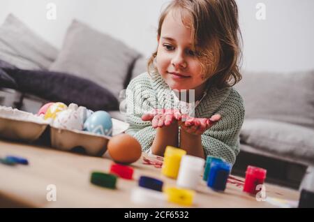 Funny kid showing hands in paints near Easter eggs in container and set of bright colors on table near sofa Stock Photo