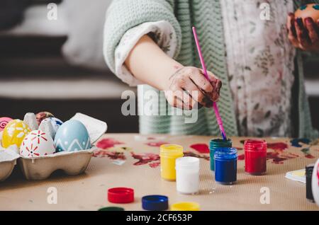 Crop kid with dirty hands in paints coloring Easter eggs in container near bright colors on table Stock Photo