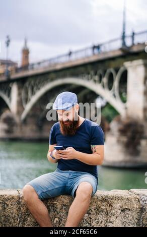 Bearded guy in casual clothing and cap sitting on rocked fence and listening voice message on mobile phone with river and big bridge on blurred background Stock Photo