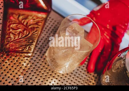 Bottle of red glass and glass full of ice cubes in hands on barman in uniform and red gloves on bar counter Stock Photo