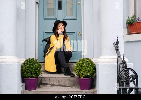 Cheerful Woman in stylish yellow coat and hat smiling and looking away while sitting near potted plants and door and answering phone call on street of London, United Kingdom Stock Photo