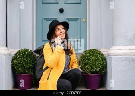 Cheerful Woman in stylish yellow coat and hat smiling and looking away while sitting near potted plants and door and answering phone call on street of London, United Kingdom Stock Photo