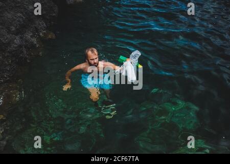 Sad man swimming in polluted water reservoir next to floating plastic bottles Stock Photo