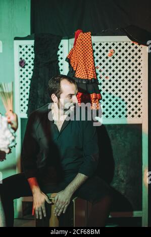 Smiling Hispanic bearded male musician playing cajon and looking away while crop anonymous female dancer with hand fan dancing flamenco on scene Stock Photo