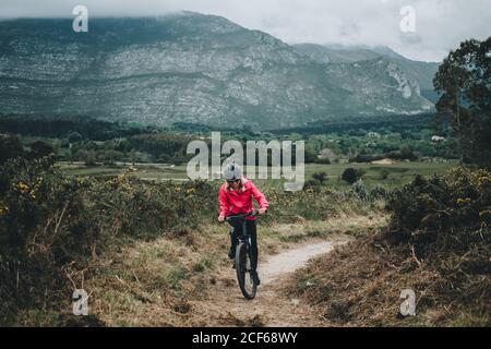 Active woman in helmet and red jacket riding bicycle on countryside road on nature background