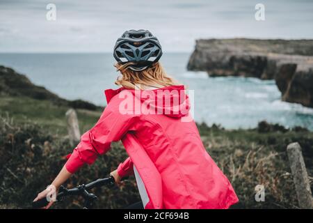 Back view of blonde woman in protective helmet riding bicycle on blurred nature background
