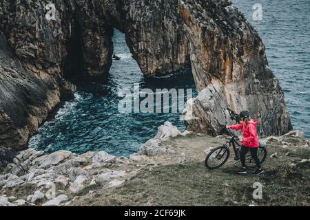 anonymous young woman with mountain bike walking on cliff on sea background