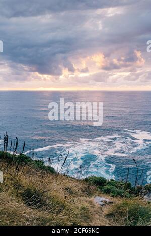 Coast with dry grass near stormy sea in cloudy evening during beautiful sunset Stock Photo