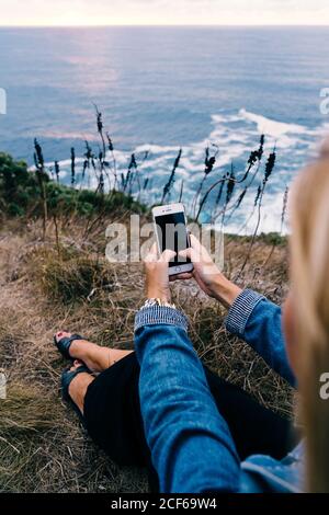 Crop hands of traveler sitting on dusky seaside and holding smartphone with black screen on blurred background Stock Photo