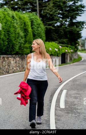 Cheerful active Woman with red jacket in hand walking along white stripe on road along green park and looking away Stock Photo