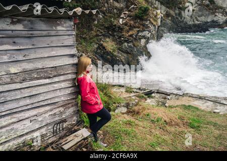 Side view of thoughtful Woman in red raincoat leaning on wooden walls of old house on seaside washed by foamy waves of sea Stock Photo