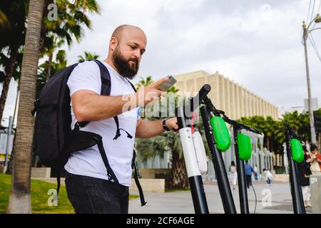 Side view of serious businessman traveling with backpack looking at mobile phone and taking scooter on parking lot for driving Stock Photo