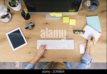 Top view of anonymous blonde caucasian Woman sitting on wooden desk writing on a notebook and working on a computer in a modern home office Stock Photo