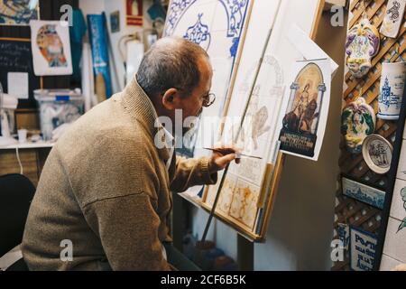 From above side view of aged focused artist in casual clothes sitting in front of paint easel and drawing patterns on ceramic tile in workshop among ceramic items Stock Photo
