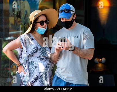 Pregnant woman, wearing protective face mask on her chin, looks on as husband, wearing face mask correctly, uses smartphone while in front of shop. Stock Photo
