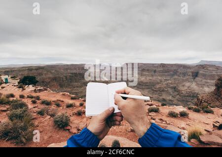 Crop man making notes in diary while having rest at edge of cliff in canyon in USA Stock Photo