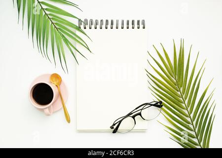 Top view of opened spiral bound notepad with blank pages placed on white background with palm leaves and trendy eyeglasses Stock Photo