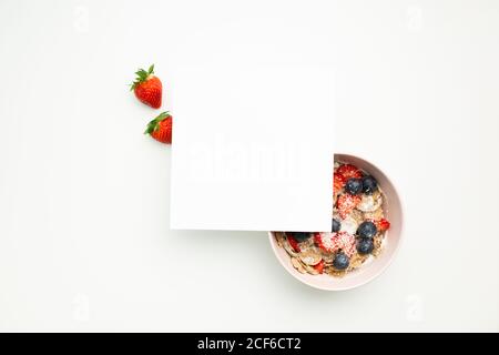 Top view of sheet of paper placed on bowl with muesli and fresh strawberries and blueberries on white desk Stock Photo