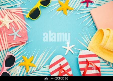 Top view composition of sunglasses and flip flops placed with beach towel and notebook on blue background with palm leaves and starfish representing summer vacation on beach Stock Photo