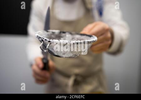 Crop man in apron standing and holding metal sieve with flour and knife on blurred background Stock Photo