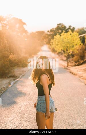Back view of content beautiful Woman in jean shorts looking over shoulder while walking along scenic countryside road in backlit Stock Photo