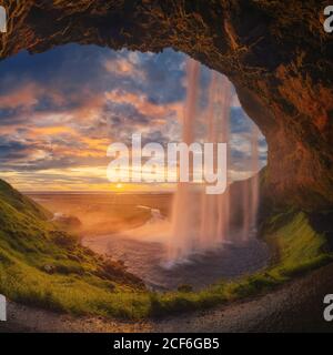 Majestic waterfall located on entrance of grassy stone cave against cloudy sundown sky on seashore in Iceland Stock Photo