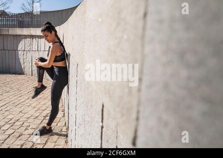 Side view of young brunette Woman in sportswear stretching before running near stone wall in park in sunny day Stock Photo