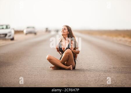 Attractive young Woman smiling and sitting on road with white stripes markup holding back back on the back with closed eyes Stock Photo
