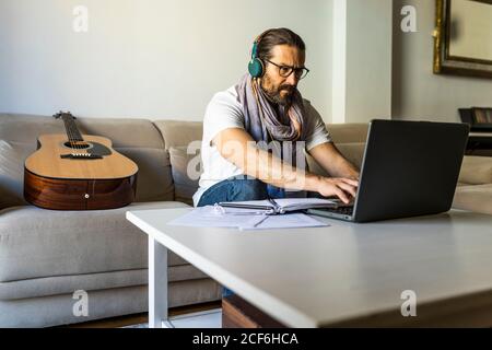 Male freelancer in casual wear and eyeglasses typing on keyboard of laptop in headphones while sitting on sofa Stock Photo