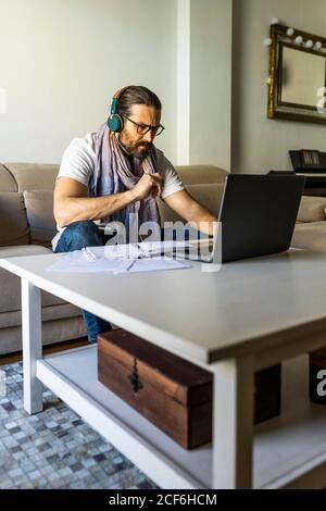 Serious bearded man in casual wear and eyeglasses watching laptop while listening music with headphones in stylish living room Stock Photo