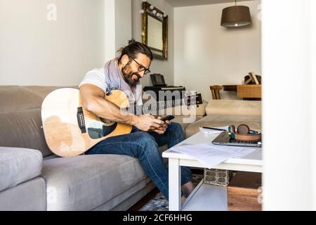 Cheerful man with guitar checking smartphone on sofa Stock Photo