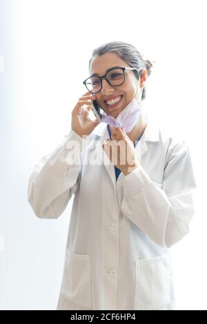 Young happy beautiful female dentist in medical gown and glasses speaking on phone and looking away isolated on white background Stock Photo