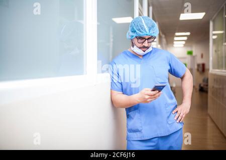 Male surgeon leaning against corridor wall while checking messages on his smart phone Stock Photo