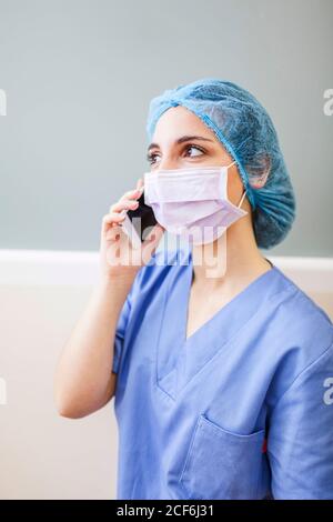 Female surgeon leaning on corridor wall while talking with her smart phone Stock Photo