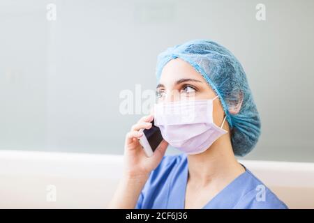 Female surgeon leaning on corridor wall while talking with her smart phone Stock Photo