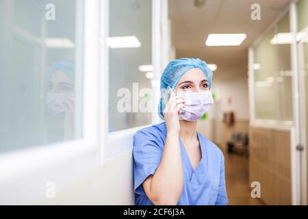 Female surgeon leaning on corridor wall while talking with her smart phone Stock Photo