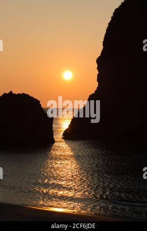 Sunlight shines between two large rocks in the sea and reflects in the calm waters as the sun sets Stock Photo