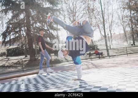 man looking at young dancer in casual wear doing handstand in park in sunny weather Stock Photo