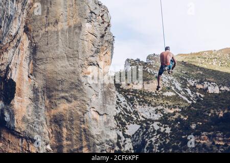 From below unrecognizable climber hanging on rope on rough cliff against blue sky Stock Photo