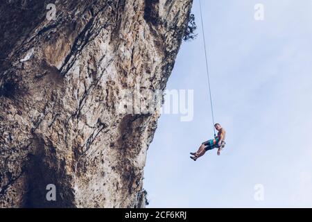 From below unrecognizable climber hanging on rope on rough cliff against blue sky Stock Photo