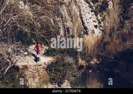 Woman with backpack and case standing on rocky coast of lake in majestic wild cliffs Stock Photo