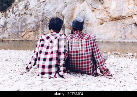 Back view of young couple sitting on rock coast of mountain river near cliff Stock Photo