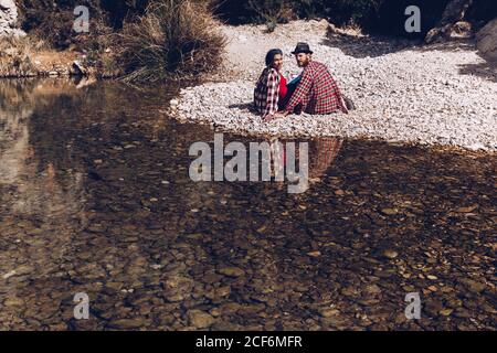 Side view of young couple sitting on rock coast of mountain river near cliff Stock Photo