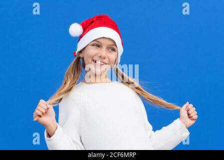 Pleased girl in white sweater and Santa hat holding pigtails smiling away isolated on blue background Stock Photo