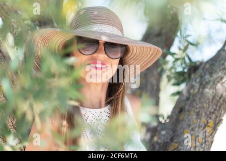 Beautiful adult Woman in stylish sunglasses and hat smiling and looking at camera while standing near tree on sunny summer day in countryside Stock Photo
