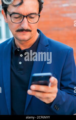 Concentrated aged male office worker with mustache in formal wear browsing mobile phone with brick wall on blurred background Stock Photo